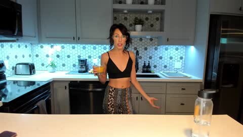 Media: Video of a slim, dark-haired woman in a black bra and leopard-print pants, holding a glass of orange juice, standing in a modern kitchen with white cabinets, black appliances, and hexagonal tile backsplash.
