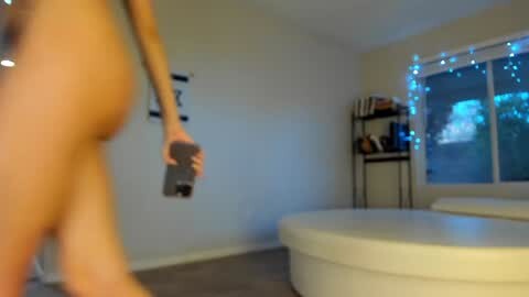 Media: Video of a naked woman with fair skin, holding a smartphone, in a modern, dimly lit living room with white walls, a round white couch, and a bookshelf.