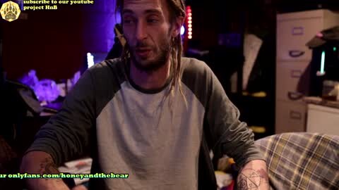 Media: A video of a bearded man with dreadlocks, wearing a grey and white long-sleeve shirt, talking into a microphone in a dimly lit room with storage cabinets and a plaid blanket.