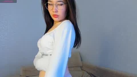 Media: Video of a young Asian woman with long dark hair, wearing glasses, a white crop top, and a \"VIXX\" belt, standing in a plain room with light blue walls.