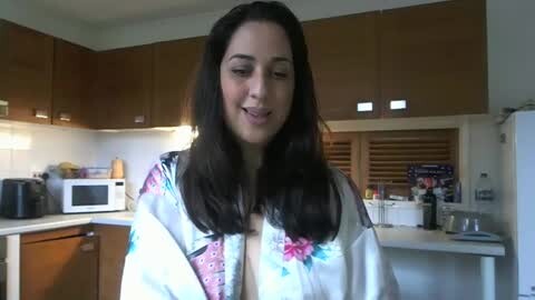 Media: Video of a smiling woman with long dark hair in a kitchen, wearing a floral robe, wooden cabinets, microwave, toaster, and white countertops.
