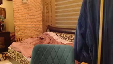 Media: A video of a cozy, rustic bedroom with a wooden headboard, beige brick wall, and a teal quilted chair. The bed is covered in a pink blanket, with a blue jacket hanging on the right.