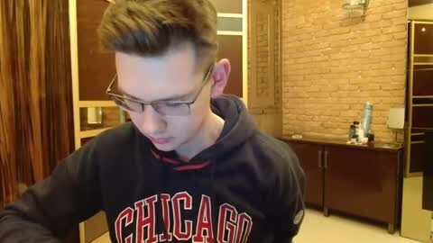 Media: Video of a young man with short brown hair and glasses, wearing a dark Chicago hoodie, in a brick-walled room with a wooden cabinet and door.