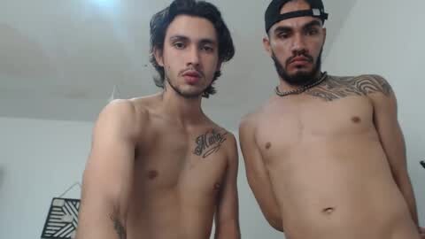 Media: Video of two shirtless, muscular men with light brown skin, one with a tattoo on his chest and a cap, the other with a beard and a necklace. Background is a minimalist white room.