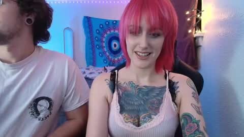 Media: A video of a smiling young woman with bright pink hair, tattoos on her chest and arms, wearing a white tank top, seated in a room with a blue tapestry and string lights.