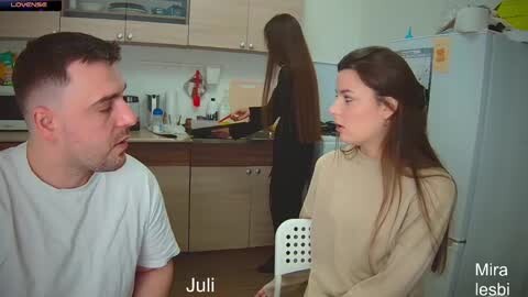 Media: Video of a white woman in beige sweater, sitting at a kitchen table, facing a man in white shirt, who looks at her. Julia and Mira are in the background.