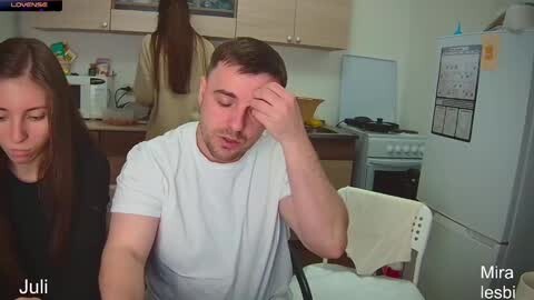 Media: Video of a white man in a white t-shirt and a white woman in a black top, both looking tired, sitting at a kitchen table in a small, cluttered kitchen.