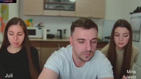 Media: Video of a sad-looking man in a white T-shirt, seated between two women in a modern kitchen with wooden cabinets, microwave, and fridge.