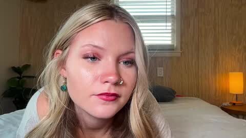 Media: Video of a fair-skinned, blonde woman with a nose ring and green earrings, wearing a white top, in a cozy, wooden-paneled bedroom with a bed, window blinds, and a lit lamp.