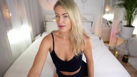 Media: Video of a blonde Caucasian woman with fair skin and medium build, wearing a black lace bra and pants, sitting on a white bed in a softly lit bedroom.