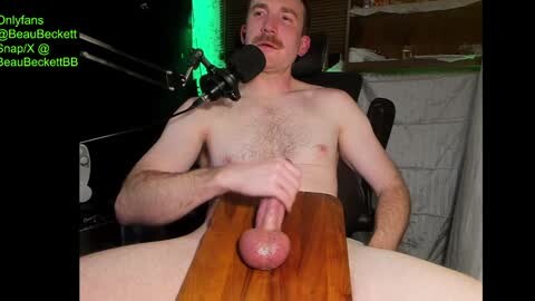 Media: A video of a shirtless, bearded white man with a circumcised, erect penis on a wooden table, wearing a headset microphone.