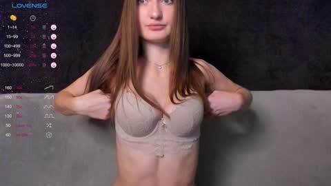 Media: A video of a young Caucasian woman with straight, long brown hair, fair skin, and medium-sized breasts, wearing a lace bra, standing against a gray wall. The background includes a streaming interface with a \"Loungese\" title and chat bubbles.