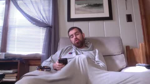 Media: Video of a man in a gray hoodie and blanket, snoozing on a beige couch in a cozy, dimly lit room with white-paneled walls, a window with blinds, and a framed painting.