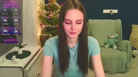 Media: Video of a young woman with long brown hair, wearing a blue shirt, sitting indoors next to a green chair, Christmas tree, and a teddy bear.