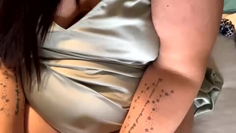 Media: Video of a person with light skin wearing a shiny, silvery-grey satin top, showing tattoos on their arms and chest.