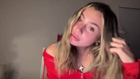 Media: Video of a young woman with long blonde hair, wearing a red zip-up hoodie, adjusting her hair in a dimly lit room with a white door and beige walls.