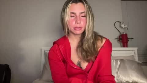 Media: Video of a blonde woman with fair skin and medium build, wearing a red hoodie, sitting on a bed with a white headboard. She has a serene expression with eyes closed. Background features a white wall and a red lamp.