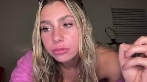 Media: A video of a young woman with long, wet blonde hair, wearing a pink robe, with a neutral expression, in a dimly lit room with closed blinds.