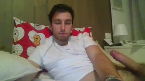 Media: Video of a young, fair-skinned man with short dark hair, wearing a white shirt, lying on a bed with red and white heart-patterned pillows. His left hand is holding a circumcised, erect penis. Background includes a wooden headboard and a nightstand with a lamp.