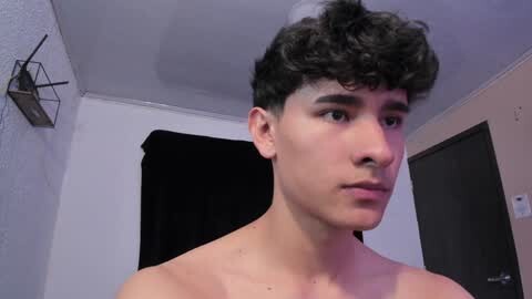 Media: Video of a young, shirtless Latino man with curly black hair, fair skin, and a neutral expression. He stands in a dimly lit room with white walls, black curtains, and a modern metal shelf.