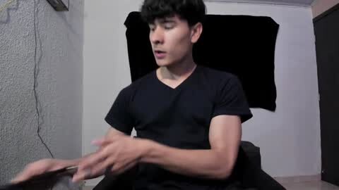 Media: Video of a young, fair-skinned, slim, dark-haired man in a black V-neck shirt, sitting in a dimly lit room with a black backdrop, hands moving in front of him.