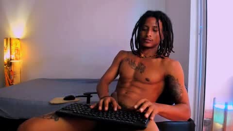 Media: Video of a shirtless, muscular black man with dreadlocks, tattoos, and a necklace, typing on a black keyboard in a dimly lit room with a lamp and blue curtains.