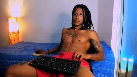 Media: Video of a shirtless, muscular Black man with dreadlocks, wearing red shorts, sitting on a blue bed, holding a black keyboard, in a dimly-lit bedroom with a lamp and blue curtains.