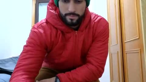Media: Video of a bearded man with a green turban, wearing a red puffy jacket, bending over, in a room with wooden doors and a bed in the background.