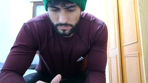 Media: Video of a man with a beard, wearing a green beanie and maroon Adidas sweatshirt, intently looking at a smartphone, indoors with light-colored walls and wooden door.