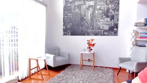 Media: Video of a minimalist, modern living room with white walls, a large black-and-white cityscape print, light gray sofa, wooden side table with orange flowers, and a plush gray rug on polished hardwood floors.
