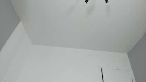 A minimalist, high-angle video of a white ceiling with a modern, black ceiling fan. The room has white walls and a door on the right side, emphasizing a clean, modern aesthetic.