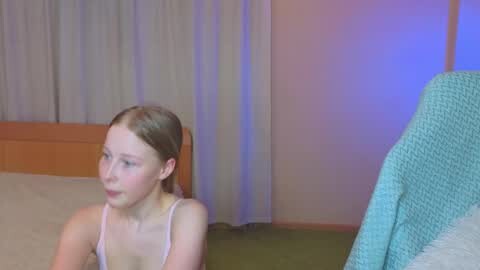 Media: Video of a young, fair-skinned girl with light brown hair, wearing a light pink tank top, sitting on a bed in a dimly lit room with a teal blanket and white curtains.