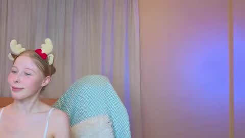 Media: Video of a fair-skinned young girl with light brown hair, wearing a red and yellow reindeer headband, a white tank top, and a blue and white blanket. She stands against beige curtains with a purple light casting a soft glow.