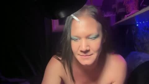 Media: Video of a blonde woman with light makeup, wearing only a green eyeshadow, with a black dog's muzzle resting on her shoulder, in a dimly lit room with purple and blue lights.
