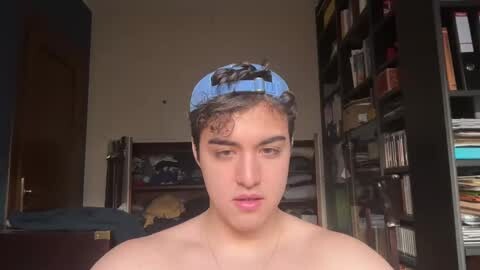 Media: Video of a shirtless young man with curly hair, wearing a blue backward cap, standing in a cluttered room with bookshelves and a partially visible bed.