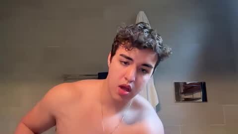 Media: Video of a young, shirtless man with curly hair, mouth open, in a bathroom with beige tiles, a towel hanging on a rack, and a showerhead.