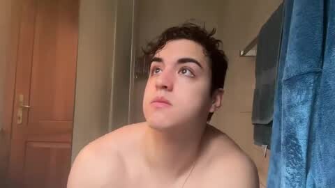 Media: Video of a young, light-skinned person with curly dark hair, wearing a silver necklace, and looking introspective in a bathroom with beige walls, wooden door, and blue towels.