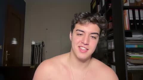 Media: Video of a shirtless young man with curly dark hair, smiling, in a dimly lit room with bookshelves and a door in the background.