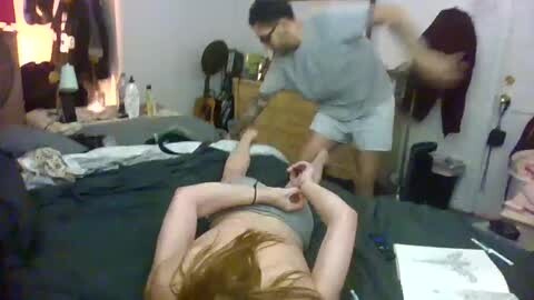 Media: A video of a young woman with fair skin and long brown hair lying on a bed in a messy room, being restrained by a man in glasses.