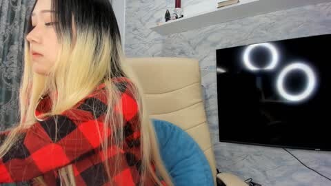 Media: Video of a young woman with long, ombre hair, wearing a red plaid shirt, sitting in a beige leather chair in a modern room with a flat-screen TV displaying a bright light pattern.