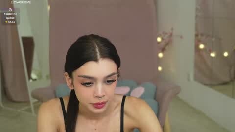 Media: Video of a young woman with dark hair, wearing a black tank top, standing in a softly lit room with a pink backdrop and a mirror.