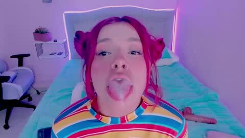Media: A video of a young woman with red hair, wearing a colorful striped shirt, lying on a bed, licking a lollipop, in a dimly lit room with purple lighting.