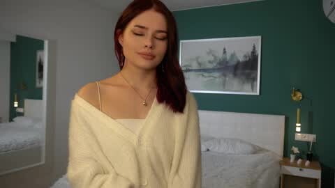 Media: Video of a young, fair-skinned woman with straight, auburn hair, wearing a white robe, standing in a modern, minimalist bedroom with a green wall, white bed, and framed forest landscape art.