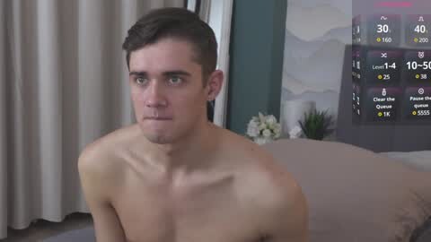 Media: A video of a shirtless, fair-skinned, young man with short brown hair and a serious expression, sitting on a bed with beige sheets, in a room with a digital TV displaying weather information.