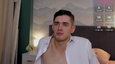 Media: Video of a young, light-skinned, attractive man with short dark hair, wearing an open white shirt, standing in a modern bedroom with a bed and abstract art wall.