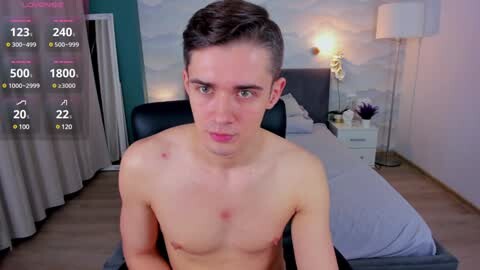 Media: Video of a young, shirtless man with short, dark hair, sitting on a bed in a modern bedroom with a white wall and nightstand, displaying live video stats in the top left.