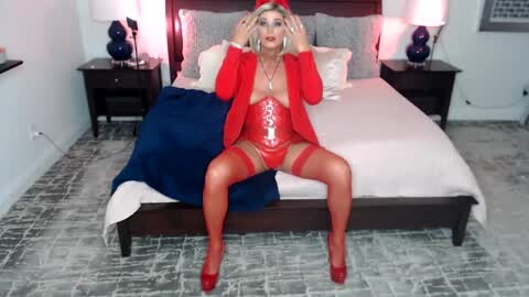 Media: Video of a curvy, blonde woman in a red corset, garter belt, and thigh-high stockings, seated provocatively on a bed with white sheets and dark wooden headboard.