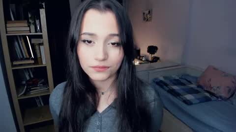 Media: Video of a young woman with long black hair, light skin, and subtle makeup, standing in a cozy bedroom with a bookshelf, blue bedspread, and a potted plant.