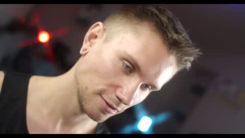 Media: Video of a fit, light-skinned man with short, spiky brown hair and blue eyes, wearing a black tank top. Background is blurred with red and blue lights.