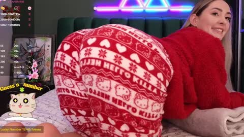 Media: Video of a smiling woman with long blonde hair, wearing a red sweater and white Hello Kitty-patterned leggings, lying on a bed, in a cozy room with neon lights and a cat photo overlay.